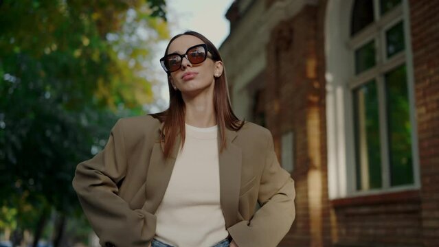 Stylish woman in jacket and trendy sunglasses posing on camera on street. Young confident female in glasses looking at camera and smile while demonstrating outfit outdoors. 