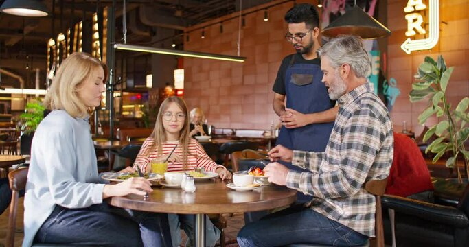 Young multiethnic waiter serving customers in restaurant. Attractive worker communicating with happy family and taking orders from them. Happy family enjoying delicious meals and dinner in cafe.