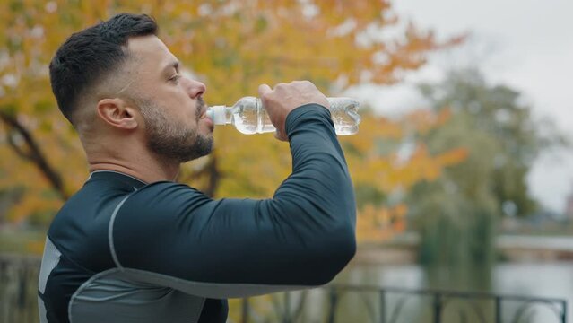 A weary male athlete, soaked in sweat, sips water from a sports bottle after a strenuous jog in a city park, showing signs of exhaustion and dehydration