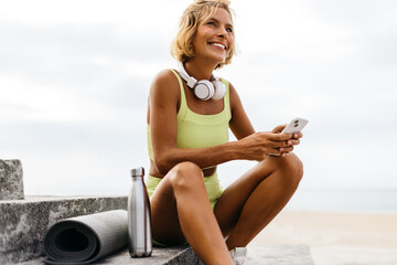 Woman sitting on beachside stairs with her yoga equipment, using her phone to browse a fitness app