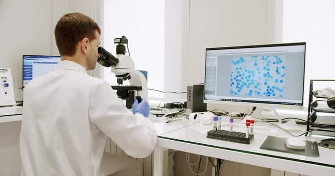 Man laboratory assistant in white uniform and rubber gloves looking blood sample trough microscope and using computer in clinic. Medical worker researching in lab with modern professional equipment