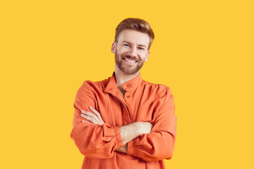 Portrait of confident smiling young bearded handsome man standing with crossed arms wearing casual bright orange summer shirt isolated on studio yellow background. Smart guy looking at the camera.