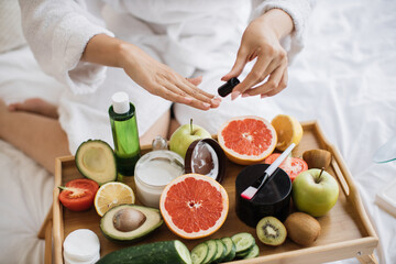 Top view of hands of multinational lady in dressing gown, paints nails while sitting near fruits and cosmetic containers on tray table on soft bed in bright room.