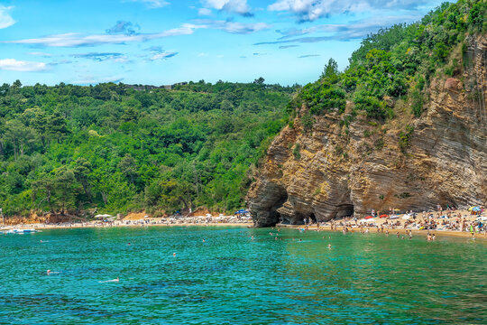 View from the Adriatic Sea to the crowded Mogren beach under the layered rocks in Budva, Montenegro. Bright turquoise green water against the coastline with many people near mountains and forest