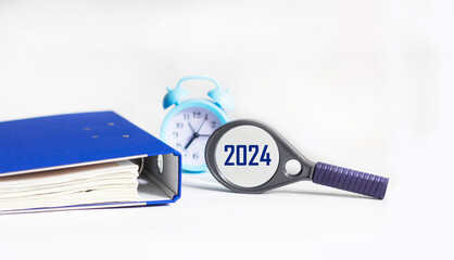 Magnifying glass with the word 2024 on a white table with a clock and a blue folder. New Year...