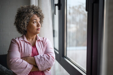 A mature woman of mixed race looking outside a large window, her expression reflecting sadness,...
