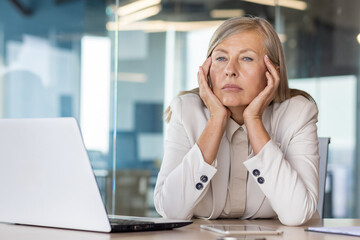 Sad unsatisfied senior woman at workplace depressed, mature business woman unhappy with achievement results at workplace unhappy sitting at table with laptop, boss thinking about problems. - obrazy, fototapety, plakaty