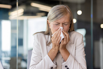 Senior gray-haired woman sick at workplace inside office, businesswoman boss sneezes has cold and...