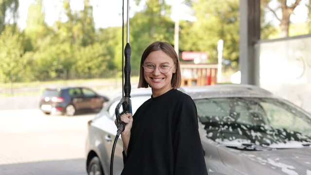 Smiling young attractive woman having fun with car washer gun, using like shot in camera. Girl is washing her vehicle at a self-service car wash. Car cleaning tools