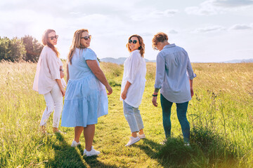 Four cheerful smiling women walking by the high green grass meadow hill, laughing and chatting with each other during a sunset walk. Woman's friendship, relations, and happiness concept image.