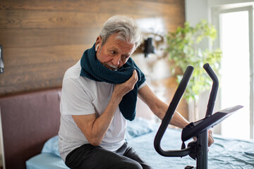 Senior man exercising on a fitness bike in the bedroom at home - Powered by Adobe