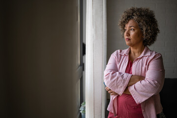 A mature woman of mixed race looking outside a large window, her expression reflecting sadness, concern, and a sense of depression