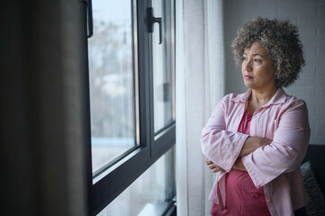 A mature woman of mixed race looking outside a large window, her expression reflecting sadness,...