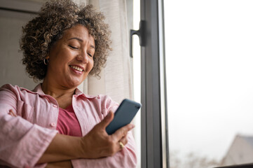 A cheerful mature woman is actively sending and receiving messages on her smartphone, spreading...