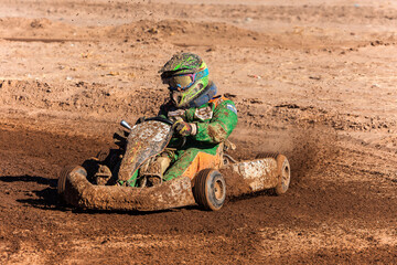 Kart driver in green overalls racing on a dirt track completely splashed with mud.