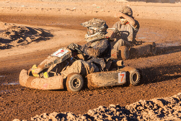 Kart drivers competing in a dirt karting, blinded by the sun and covered in mud.