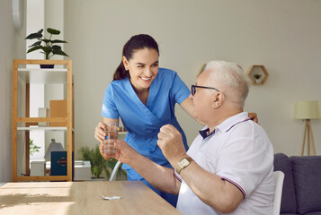 Friendly young female nurse giving medication and glass of water to senior man in nursing home....