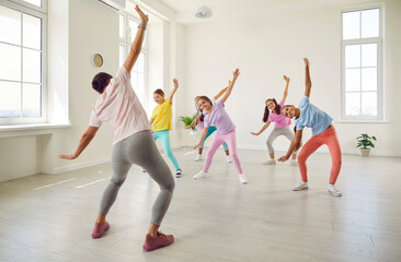 Obraz premium Female young choreographer showing her students girls stretching exercises in dance studio. Happy kids doing dance workout. Children training in choreography class, kids sport concept.