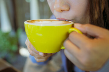 Closeup image of a cup of coffee with heart shape latte art, young woman drinking coffee in coffee shop. Soft focus.