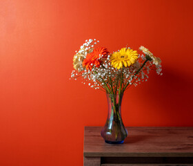Bouquet of gerberas and gypsophila in blue glass vase on wooden table against orange wall.