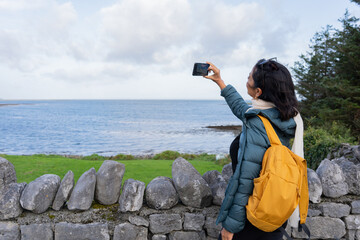 Latin woman traveler with a yellow backpack next to a gray stone wall taking a photo in a beautiful landscape with the sea in the background and the green field in ireland - obrazy, fototapety, plakaty