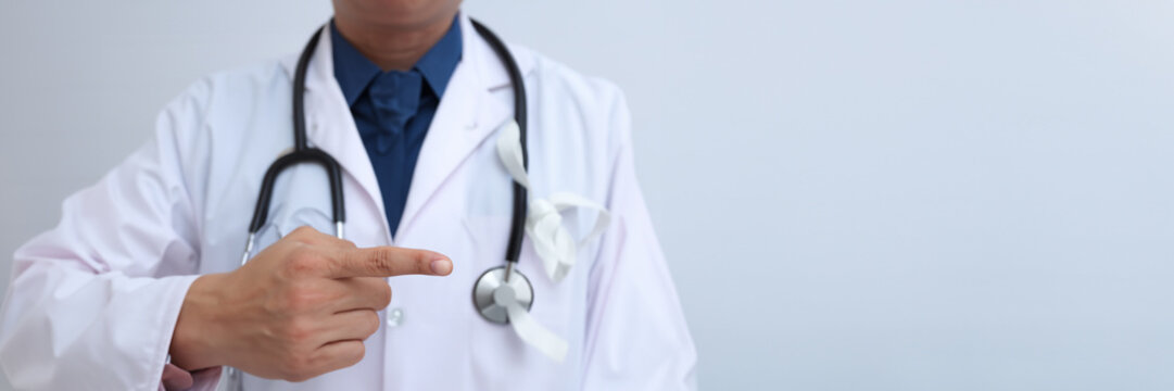 Handsome Bearded Male Medical Physician In White Lab Coat Points Finger Toward Upper Right Corner Of Frame On White Background With Copy Space