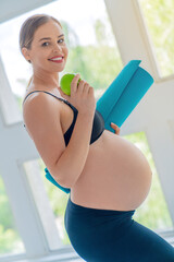 Beautiful pregnant woman standing with sport blue mat and eating apple, only healthy eating