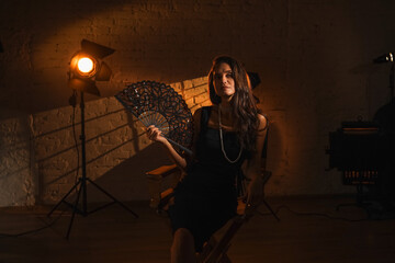 Portrait of attractive female actress. Brunette woman in black dress with hairstyle and pearls sitting on directors chair and holding lace fan.