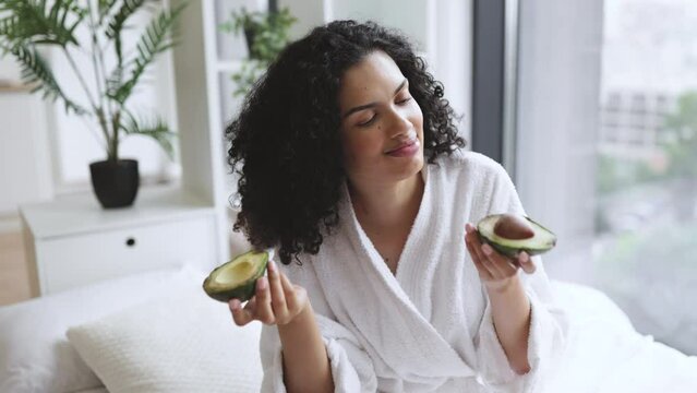 Beautiful calm multiracial female in robe sniffs avocado halves in hand near face in bedroom interior. Young African American curly woman appreciating green avocado for great skin benefits.