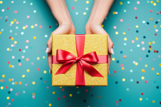 Close-up Of A Woman's Hands Holding A Small Gift Wrapped In A Red Ribbon, Against A Bright Background With Colored Confetti. View From Above