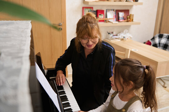 Beautiful mature woman, pianist teacher giving music lesson to a smart child girl, teaching her playing on grand piano