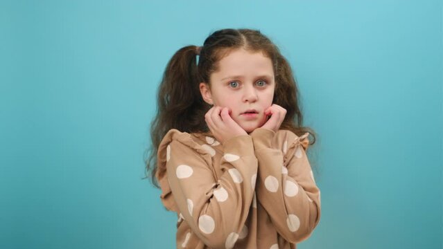 Portrait of afraid preteen caucasian girl child covering mouth, hiding back from scary content, looking at camera, posing isolated over plain blue color background wall in studio. Kid fears concept