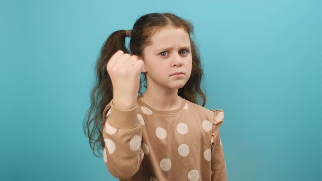 Portrait of aggressive angry preteen girl kid trying to fight at camera, shaking fist, boxing with expression, punishment, disappointment, posing isolated over blue color background wall in studio