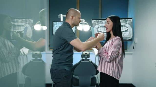 Side view of doctor and patient standing in fully eqipped illuminated office near dentist chair choosing dentures. Dental expert uses samples of implants. High quality 4k footage
