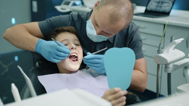 Brave boy sitting in dentist chair with napkin on chest, mouth wide open, dental expert in mask and medical gloves uses tooth scraper, concave mirror. High quality 4k footage