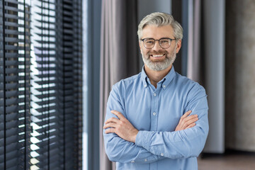 Portrait of mature successful businessman boss, senior gray-haired man inside office smiling looking at camera with crossed arms, experienced financier banker accountant in shirt.