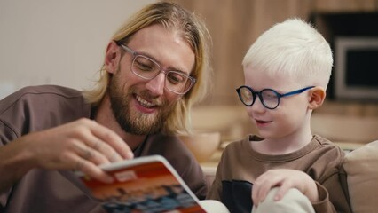 Close-up: a blond man with a beard and glasses helps his little albino son with white hair in blue round glasses to do his homework and reads for him what is written in a notebook. Sitting on the sofa