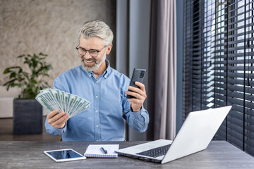 Fototapeta premium Happy successful financier investor inside office at workplace, senior gray haired businessman holding phone and money cash US dollars in hands, boss celebrating victory and successful achievement.