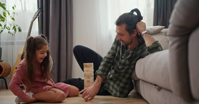 A little brunette girl in a pink dress plays with her father a brunette man in a checkered green shirt in the board game Jenga while sitting on the floor leaning on a gray sofa in a modern apartment