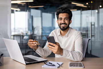 Portrait of a smiling young man working in the office, sitting at a desk with a laptop, holding a...