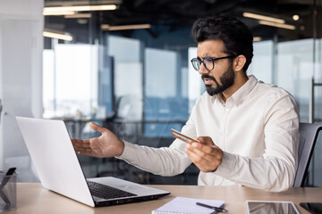 Frustrated and worried young muslim man sitting in office and looking at laptop screen, holding credit card in hand and waving hands in confusion