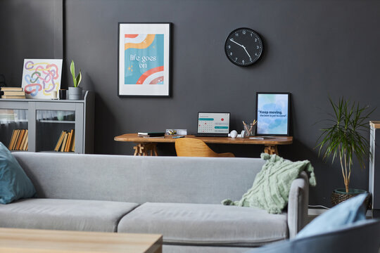Wide shot of cozy flat interior with home office area including big sofa, wooden table and motivational posters at dark-gray wall