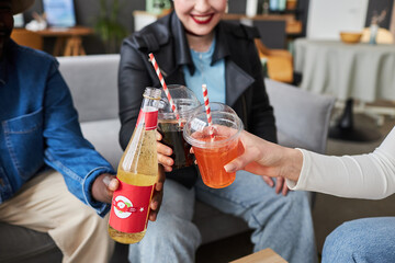 Close up shot of fizzy drinks in plastic cups and bottle in hands of diverse friends toasting at home - obrazy, fototapety, plakaty