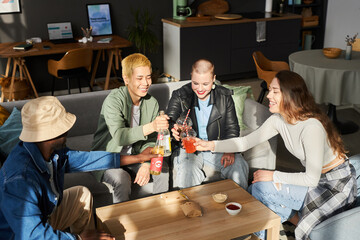 Medium long shot of four happy multi-ethnic young friends toasting with fizzy drinks sitting at...