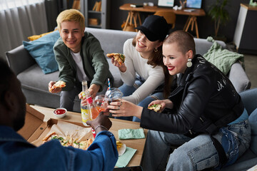 High angle view at diverse group of smiling young adults in living room holding slices of pizza and toasting with soda - obrazy, fototapety, plakaty