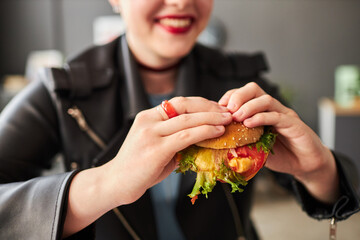 Close up shot of big burger with vegetables in hands of defocused smiling woman in leather jacket