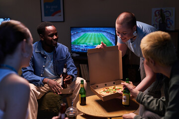 Medium full shot of diverse young friends at home drinking and watching football, smiling young girl putting cardboard box with pizza on small table