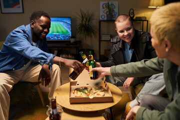 Medium full shot of three diverse young adults toasting bottles of beer while watching football and eating pizza at home