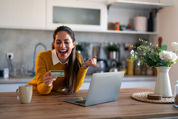 Joyful businesswoman feeling excited holding credit card sitting at desk in the kitchen with laptop...