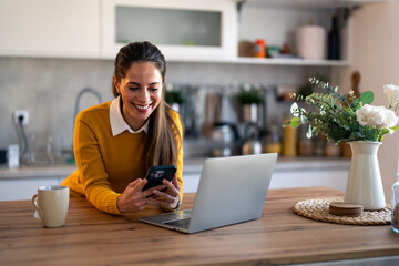 Smiling female freelancer scrolling social media on cellphone at desk in home office. Young woman digital nomad using mobile phone and laptop computer indoors.
